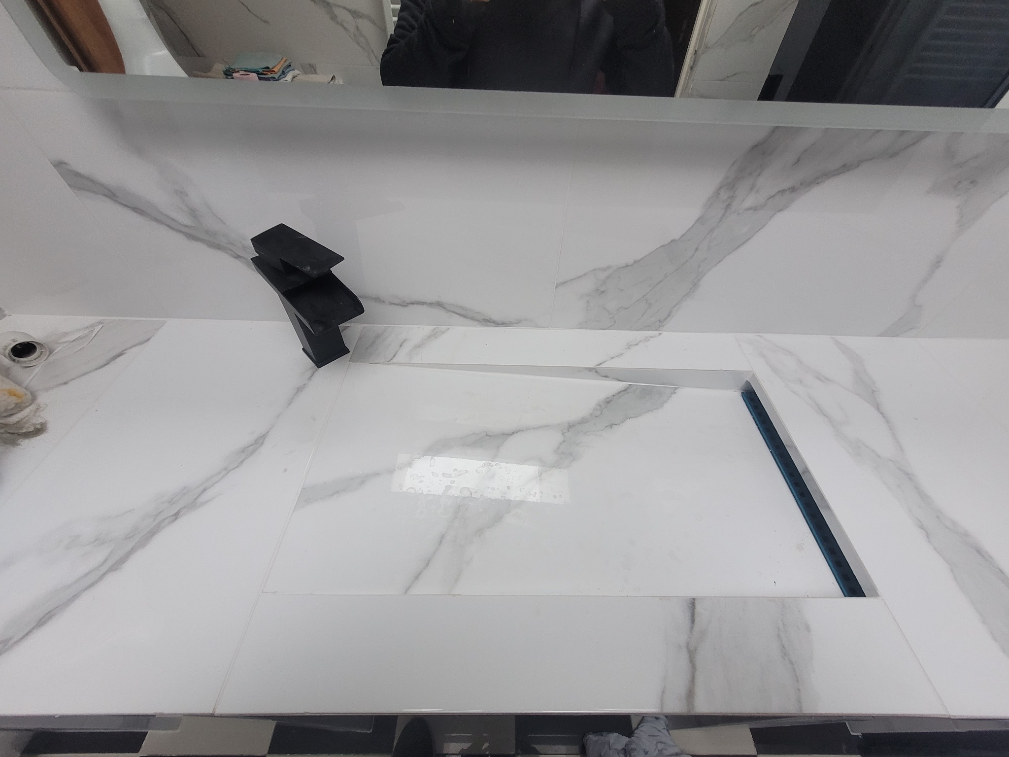 Biała umywalka z marmurowym wzorem i czarną, nowoczesną baterią, widoczna w lustrze osoba.