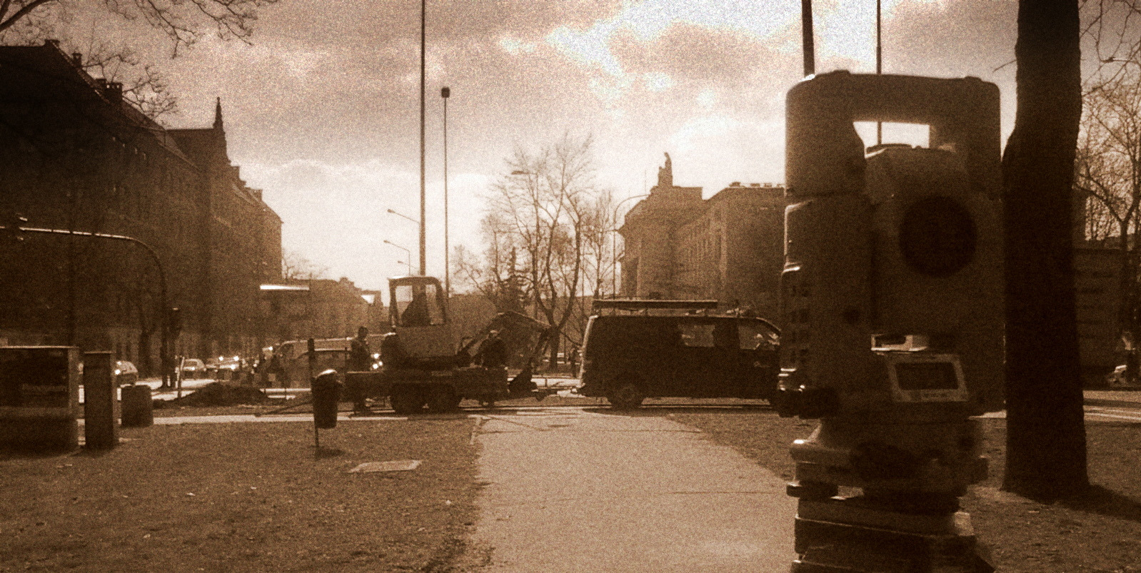 Tachimetr ustawiony na tle miejskiego krajobrazu z pracującą koparką i samochodem dostawczym, w sepii.