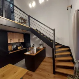 Wnętrze mieszkania z drewnianymi schodami spiralnymi z czarną metalową balustradą, drewniana podłoga, widoczna kuchnia z drewnianymi szafkami i wyspą, oświetlenie wiszące z żar&oacute;wkami Edison.