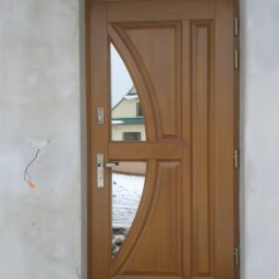 FRANCISZEK MIKOŁAJCZYK ZAKŁAD STOLARSKI - Brązowe drzwi wejściowe z łukowym przeszkleniem, widoczne niedokończone wykończenie ściany i fragment zaśnieżonego podwórka w odbiciu.