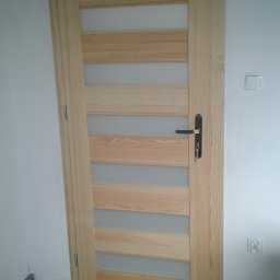FRANCISZEK MIKOŁAJCZYK ZAKŁAD STOLARSKI - Jasne, drewniane drzwi wewnętrzne z przeszkleniami ułożonymi horyzontalnie, widoczna klamka z kluczem, biała ściana z włącznikiem i gniazdkiem elektrycznym, szara podłoga.