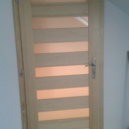 FRANCISZEK MIKOŁAJCZYK ZAKŁAD STOLARSKI - Jasne, drewniane drzwi wewnętrzne z poziomymi przeszkleniami, zamontowane w skośnej ścianie poddasza, widoczne zawiasy i klamka.