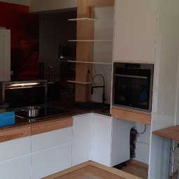 FRANCISZEK MIKOŁAJCZYK ZAKŁAD STOLARSKI - Wnętrze kuchni w trakcie montażu: białe szafki z drewnianymi akcentami, płyta indukcyjna, piekarnik i drewniane półki na tle czerwonej ściany z motywem palmy.