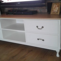 FRANCISZEK MIKOŁAJCZYK ZAKŁAD STOLARSKI - Biała komoda RTV z drewnianym blatem, widoczne dwie półki i cztery szuflady z ozdobnymi uchwytami, na blacie telewizor, ramki ze zdjęciami i dekoracje, komoda stoi na drewnianej podłodze.