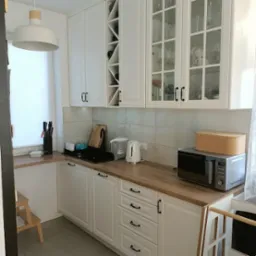 Jasna kuchnia z białymi szafkami, drewnianym blatem i szklanymi frontami g&oacute;rnych szafek, widoczna p&oacute;łka na wino i drobne akcesoria kuchenne.