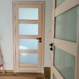 FRANCISZEK MIKOŁAJCZYK ZAKŁAD STOLARSKI - Dwie drewniane drzwi wewnętrzne z matowymi szybami i czarnymi klamkami, widoczne na tle białej ściany i drewnianej podłogi.