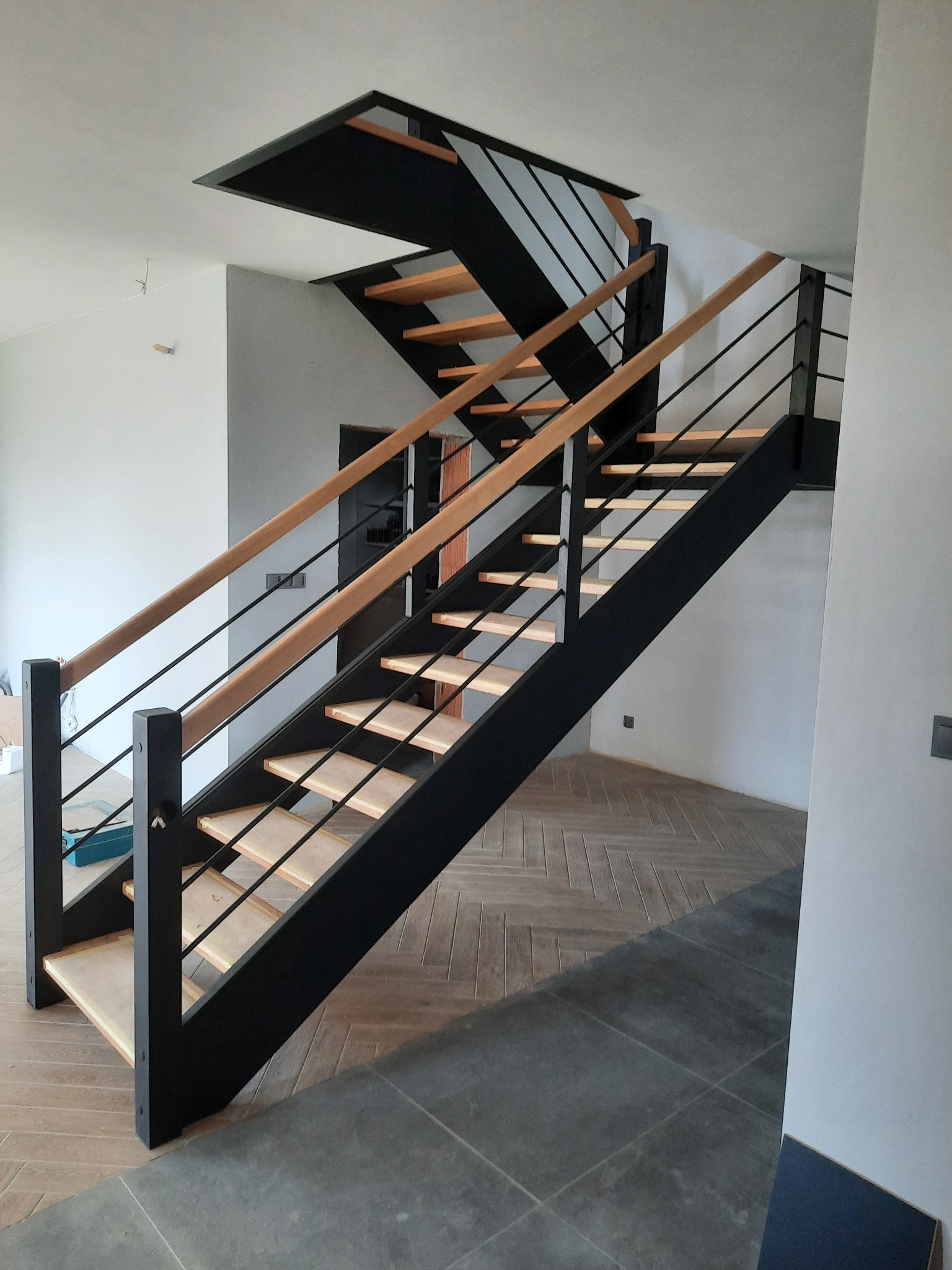 Nowoczesne schody z czarną konstrukcją i jasnymi drewnianymi stopniami, drewniana poręcz, widok z dołu na tle surowych ścian i zróżnicowanej podłogi.