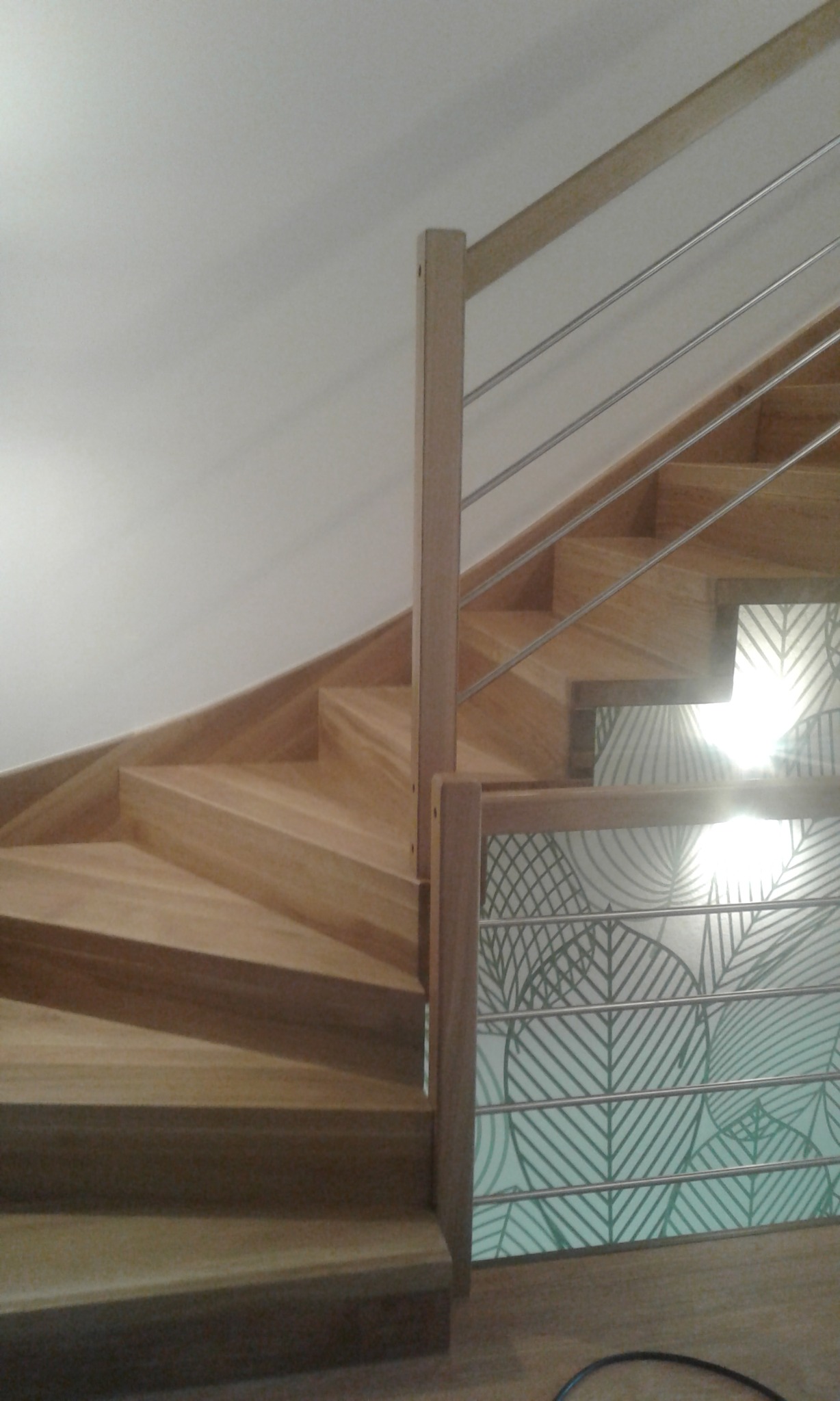 Jasne drewniane schody z metalowymi poręczami i dekoracyjnym panelem z motywem liści na ścianie.