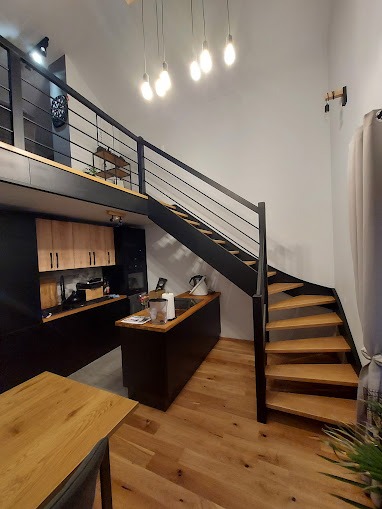 Wnętrze mieszkania z drewnianymi schodami spiralnymi z czarną metalową balustradą, drewniana podłoga, widoczna kuchnia z drewnianymi szafkami i wyspą, oświetlenie wiszące z żarówkami Edison.