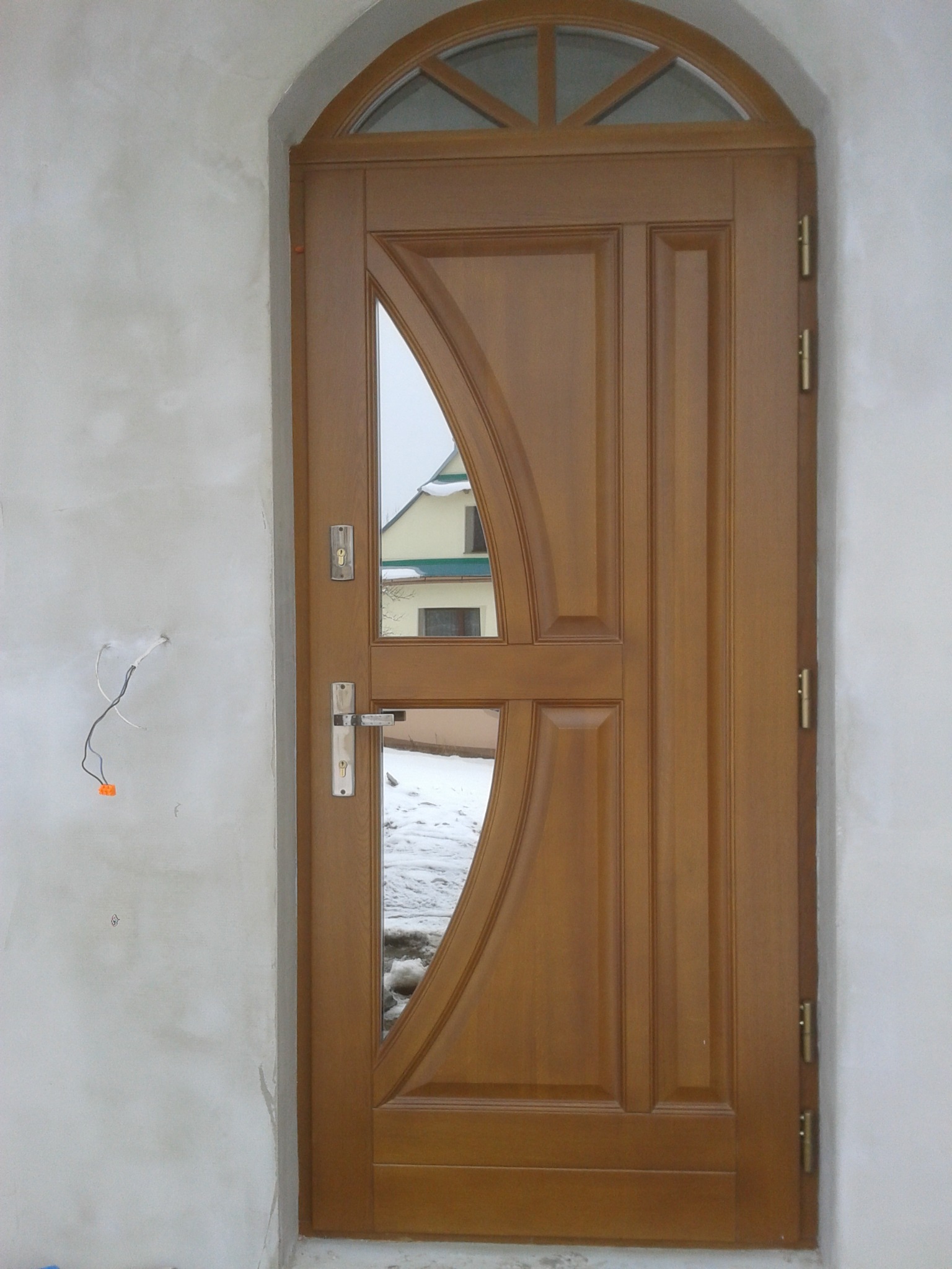 Brązowe drzwi wejściowe z łukowym przeszkleniem, widoczne niedokończone wykończenie ściany i fragment zaśnieżonego podwórka w odbiciu.
