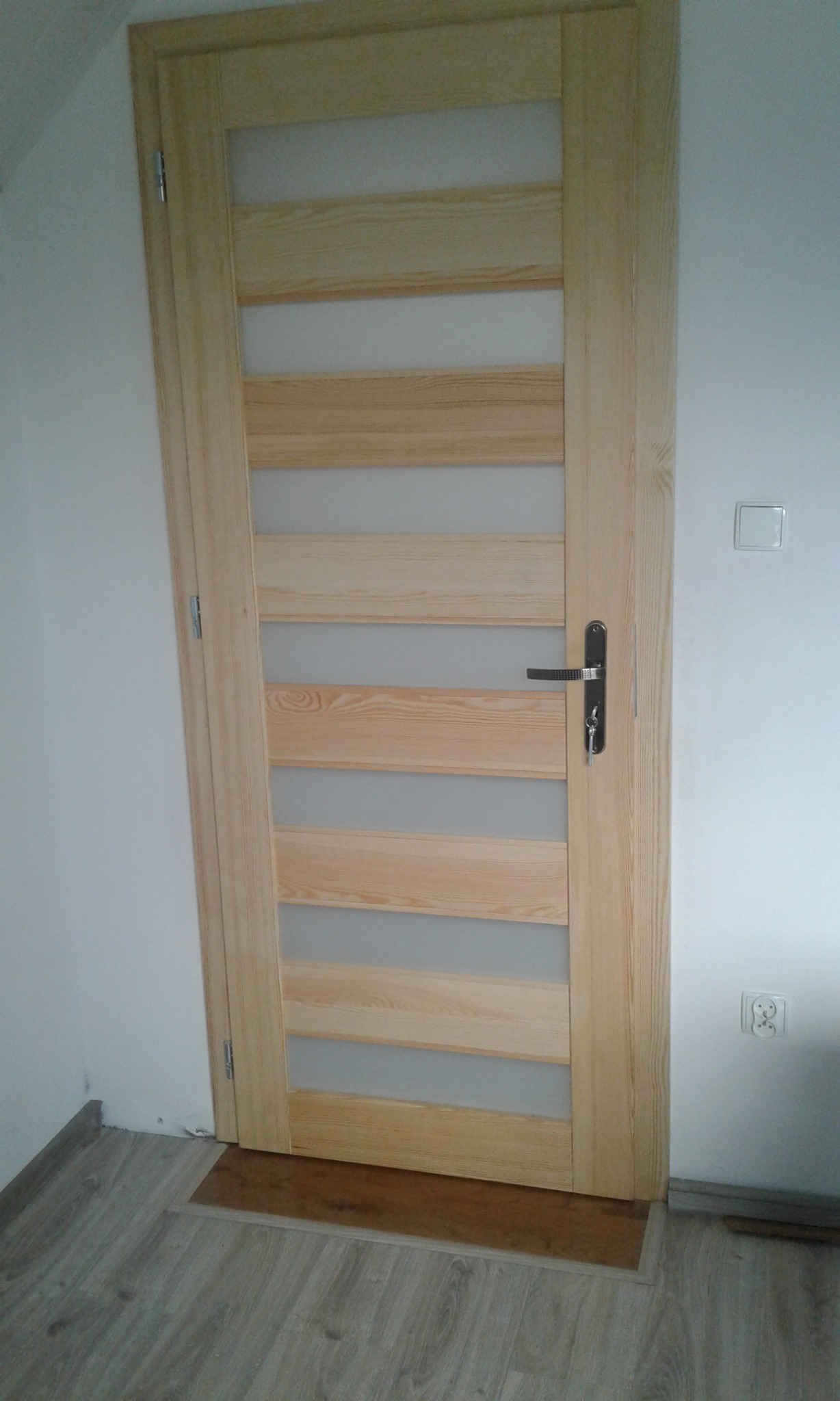 Jasne, drewniane drzwi wewnętrzne z przeszkleniami ułożonymi horyzontalnie, widoczna klamka z kluczem, biała ściana z włącznikiem i gniazdkiem elektrycznym, szara podłoga.