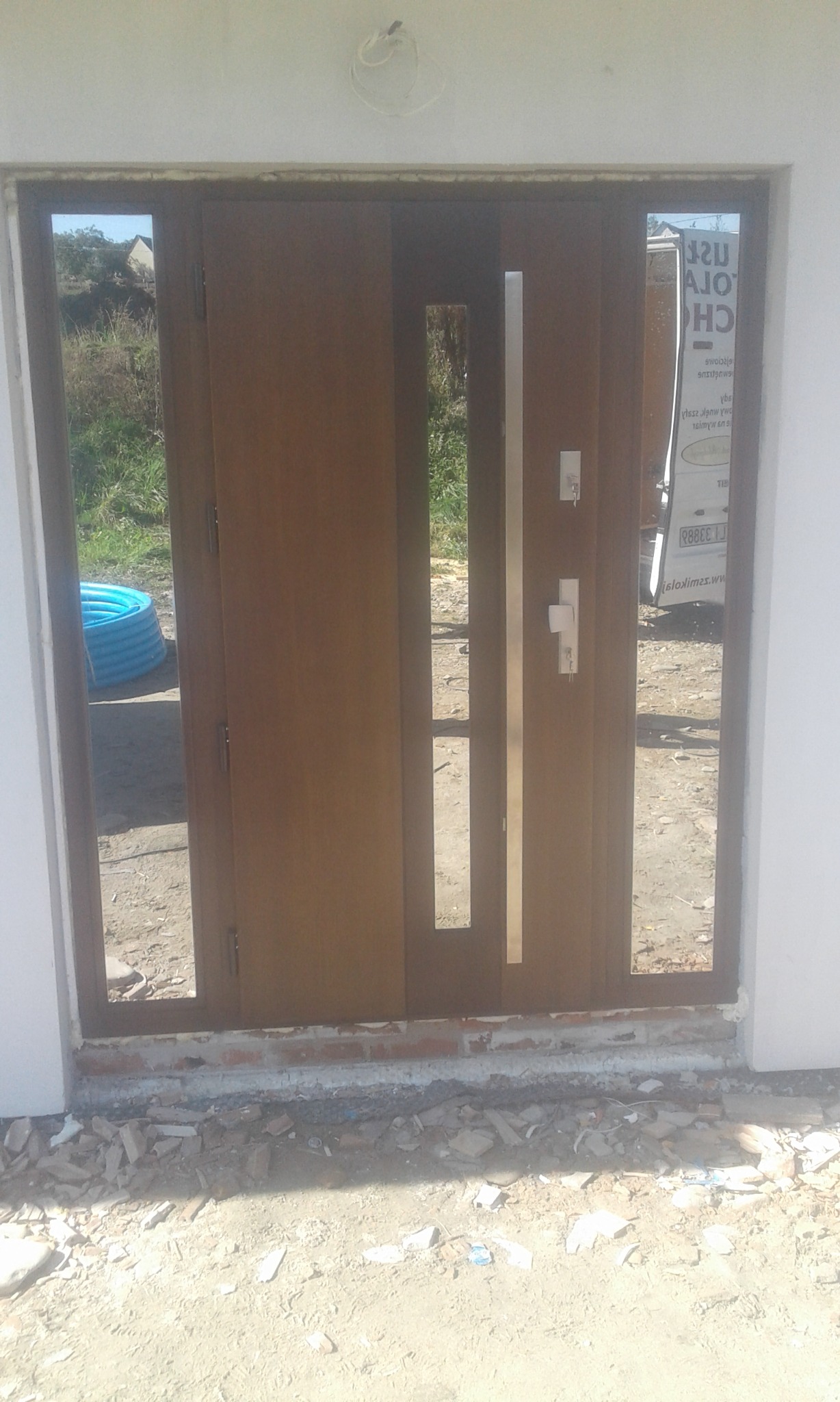 Nowe, brązowe drzwi wejściowe z przeszkleniami bocznymi i metalową listwą, widoczne ślady prac murarskich i gruz przed wejściem, w tle częściowo widoczny samochód dostawczy z reklamą firmy.