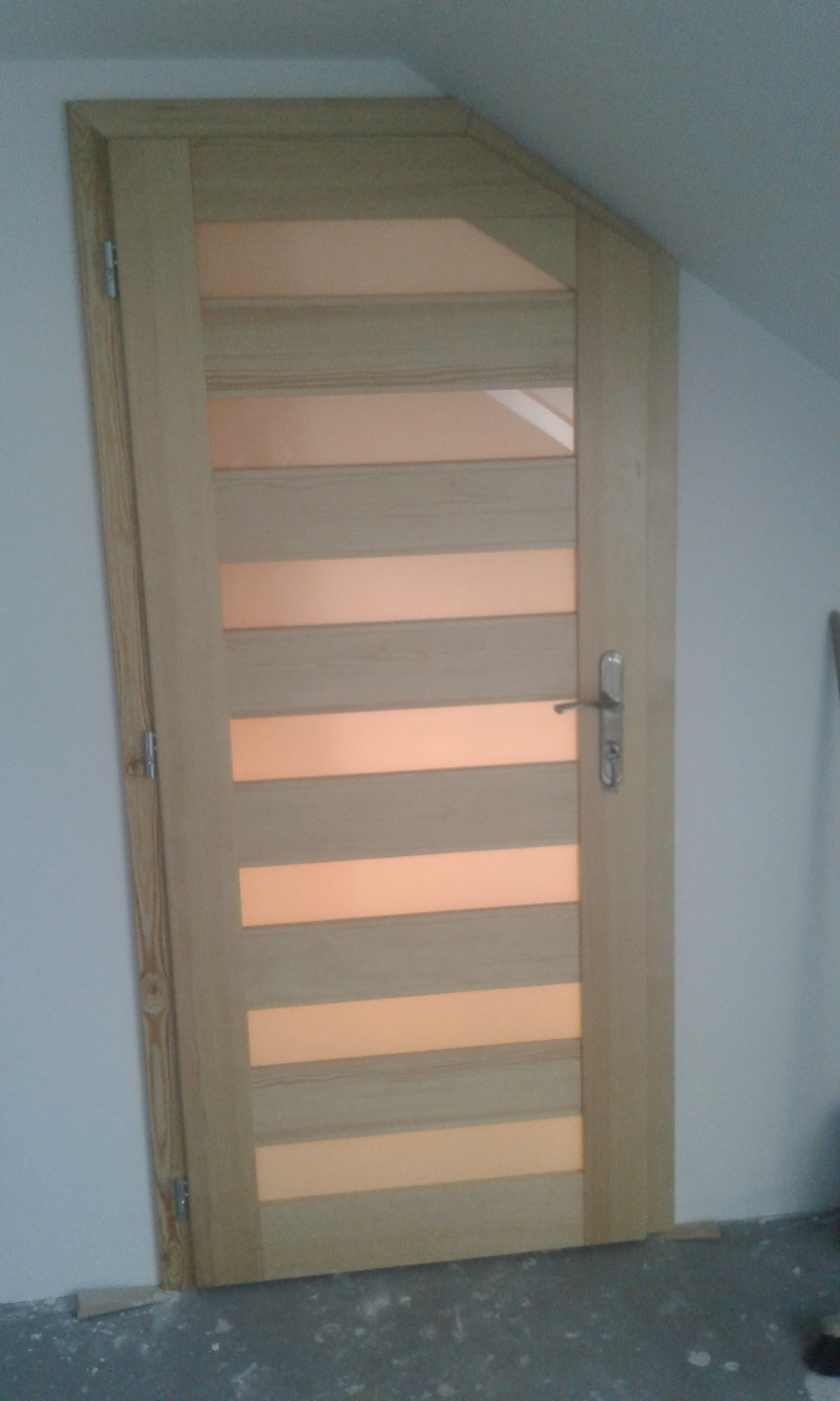 Jasne, drewniane drzwi wewnętrzne z poziomymi przeszkleniami, zamontowane w skośnej ścianie poddasza, widoczne zawiasy i klamka.