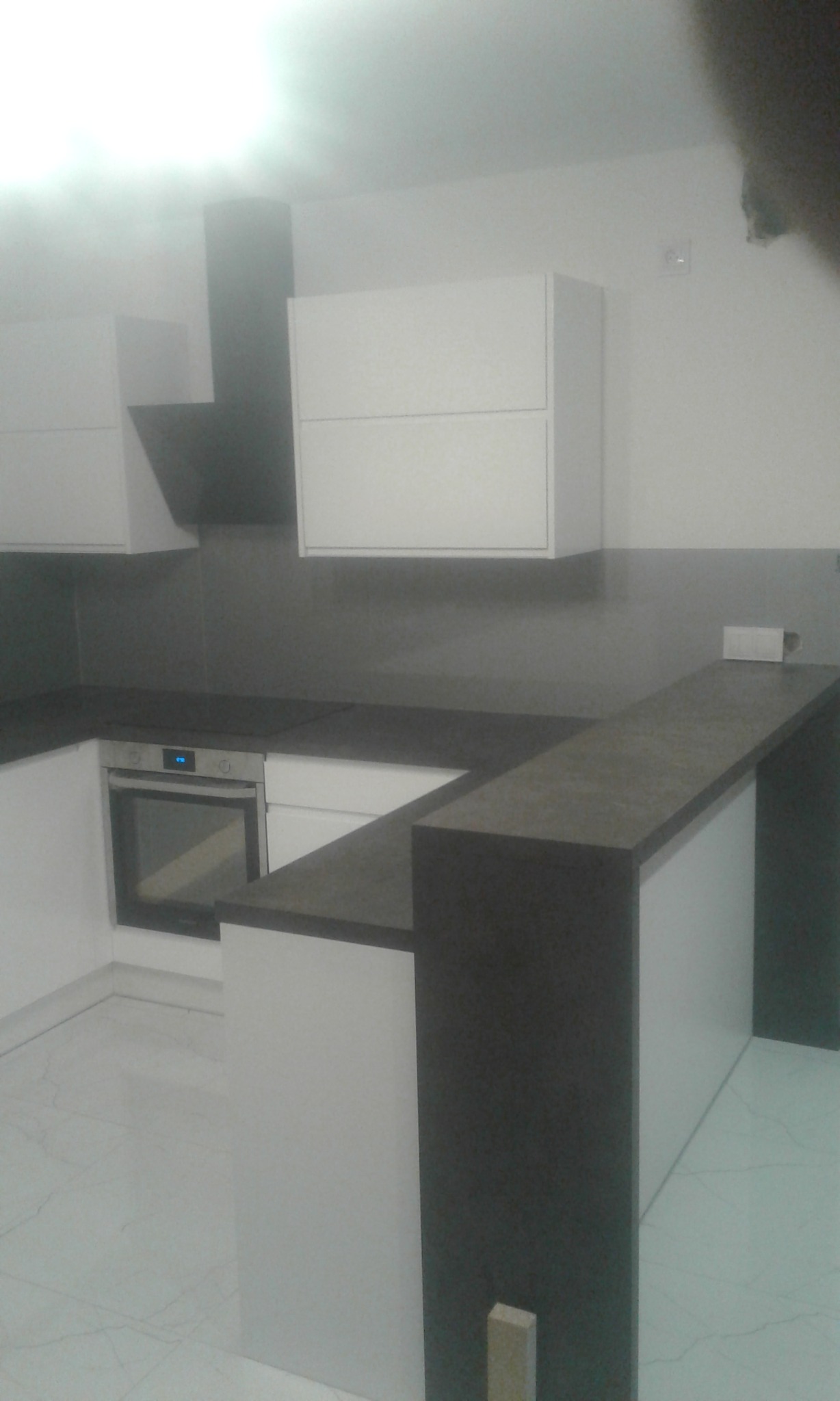 Nowoczesna kuchnia z białymi szafkami i ciemnym blatem, widok na piekarnik i okap, marmurowa podłoga.