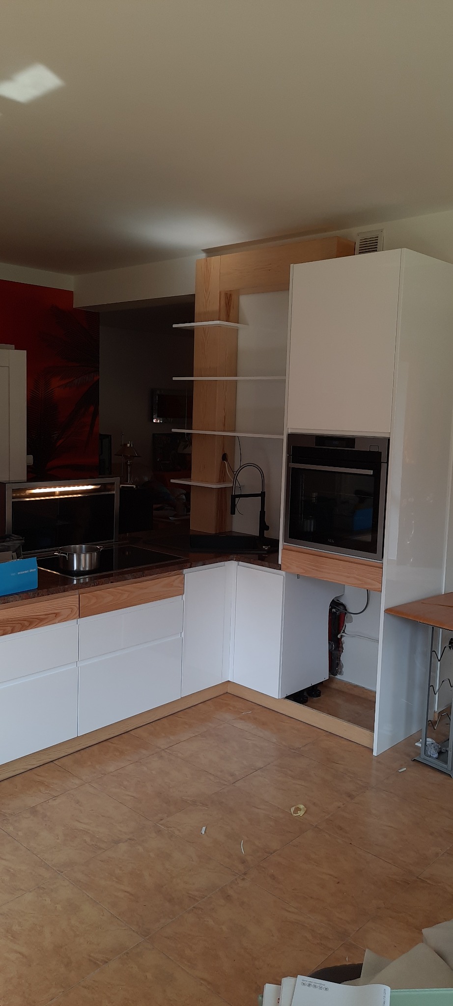 Wnętrze kuchni w trakcie montażu: białe szafki z drewnianymi akcentami, płyta indukcyjna, piekarnik i drewniane półki na tle czerwonej ściany z motywem palmy.