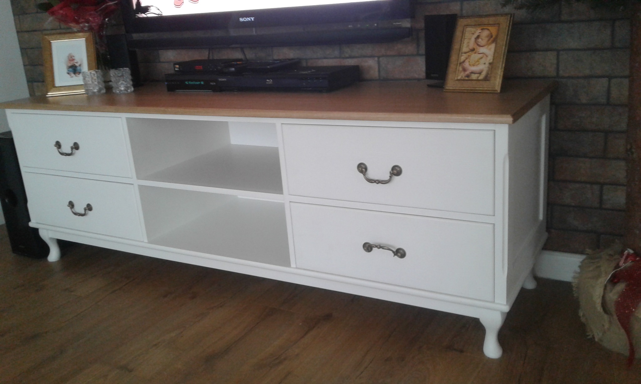 Biała komoda RTV z drewnianym blatem, widoczne dwie półki i cztery szuflady z ozdobnymi uchwytami, na blacie telewizor, ramki ze zdjęciami i dekoracje, komoda stoi na drewnianej podłodze.