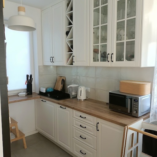 Jasna kuchnia z białymi szafkami, drewnianym blatem i szklanymi frontami górnych szafek, widoczna półka na wino i drobne akcesoria kuchenne.