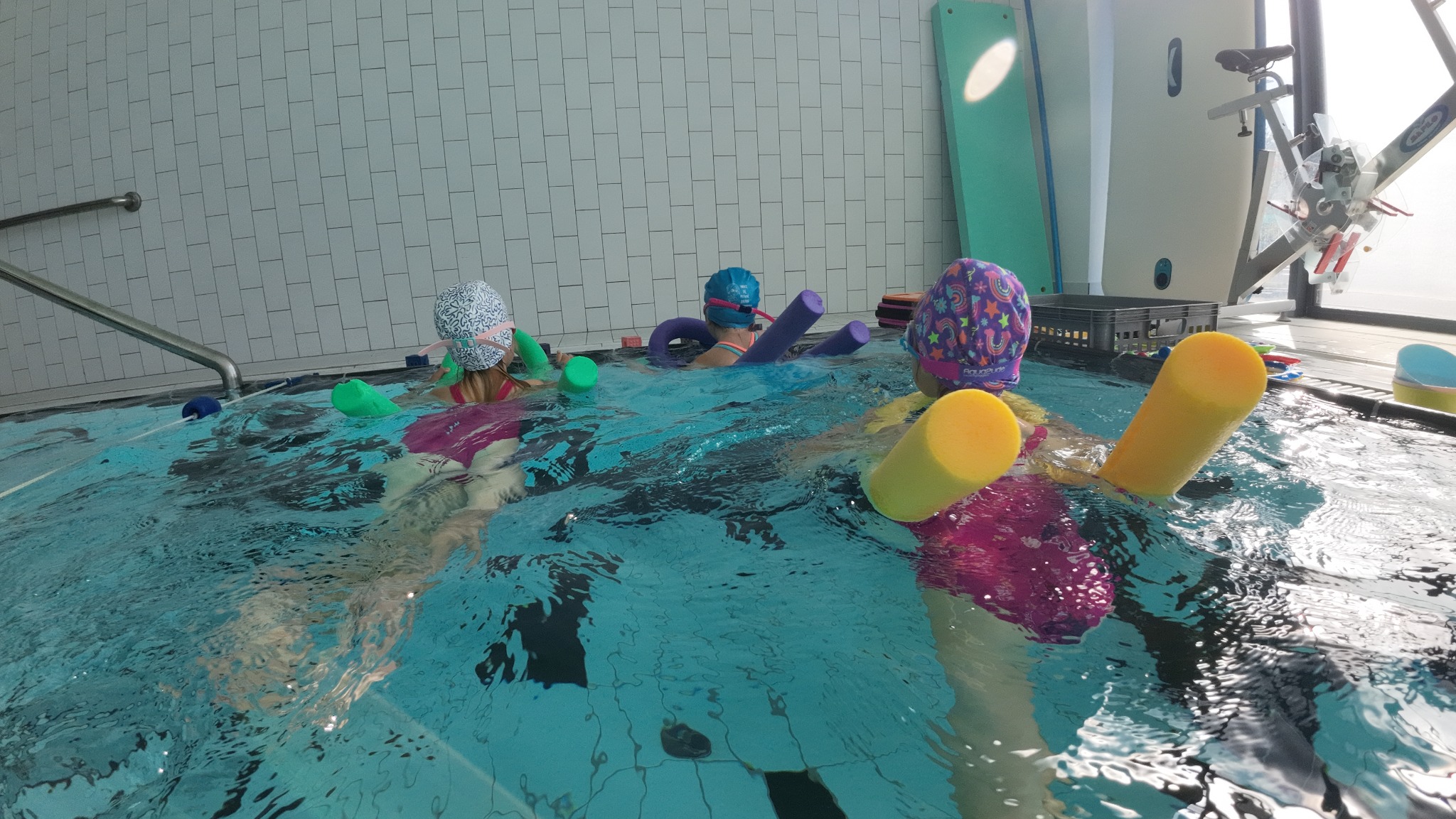Trójka dzieci w czepkach i z makaronami pływackimi w basenie z białymi kafelkami w tle; widoczny sprzęt do ćwiczeń na brzegu basenu.