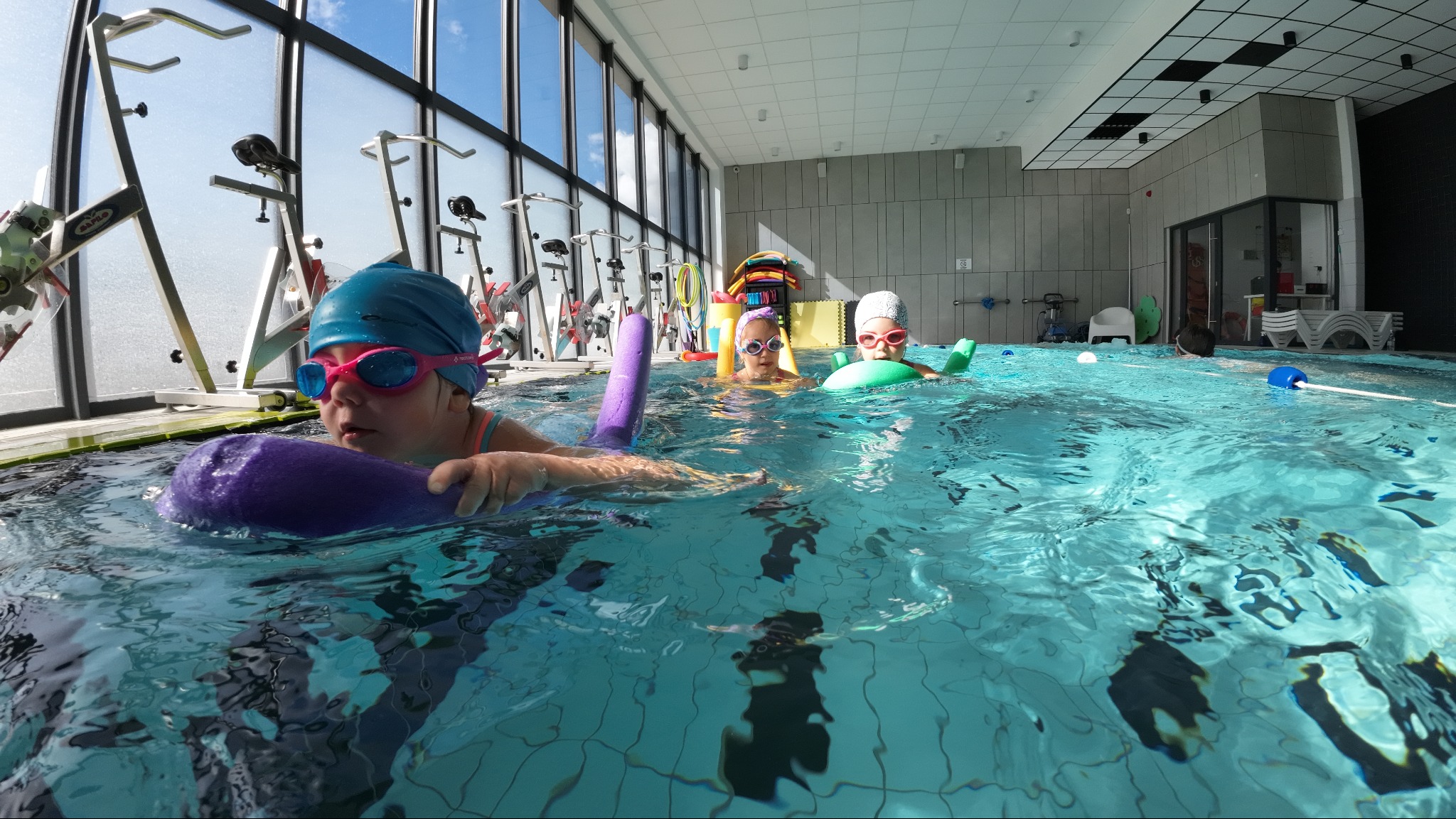 Dziecko w niebieskim czepku i różowych okularach pływa w basenie trzymając fioletowy makaron, w tle inne dzieci i sprzęt do ćwiczeń w wodzie.