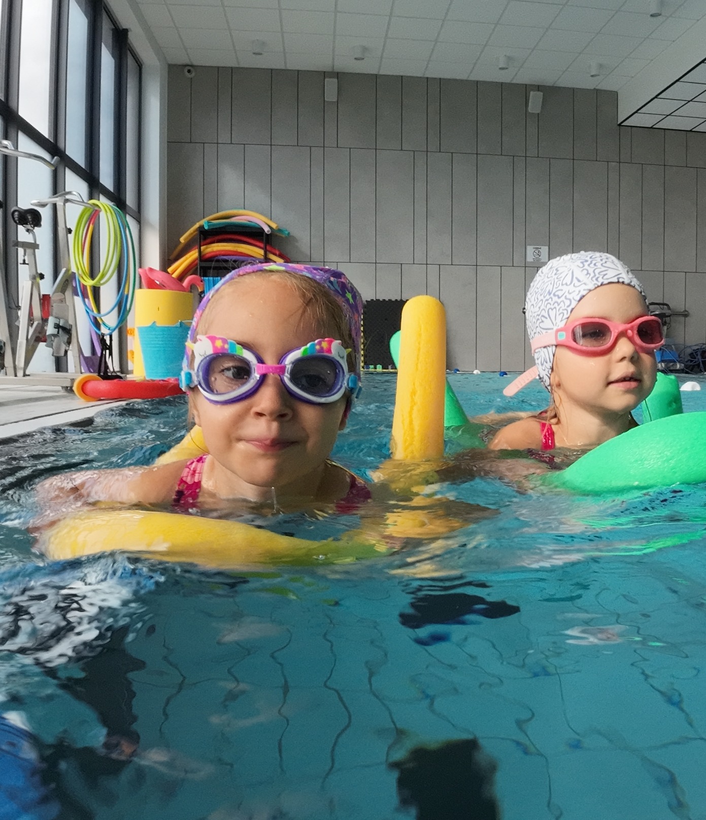 Dwie dziewczynki w czepkach i okularach pływackich, unoszące się na wodzie w basenie z makaronami piankowymi, w tle widoczne akcesoria do nauki pływania i okno.