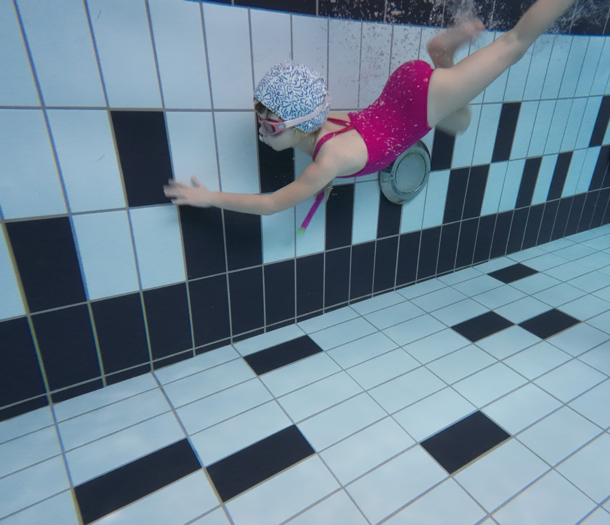 Dziewczynka w różowym kostiumie i czepku z niebieskim wzorem pływa pod wodą w basenie z biało-czarnymi płytkami.