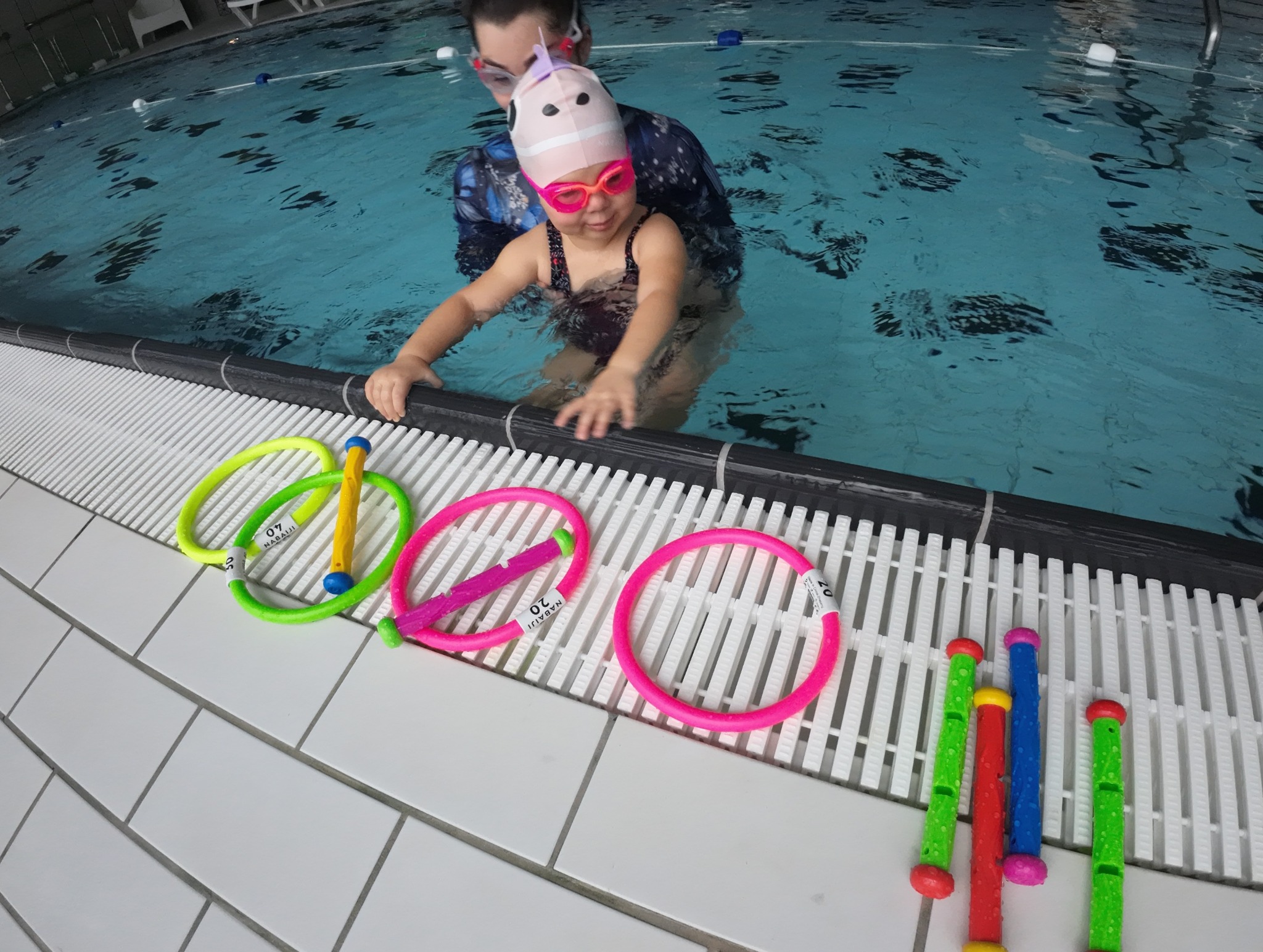 Instruktor z dzieckiem w różowym czepku i okularach w basenie, przy krawędzi leżą kolorowe obręcze i zabawki do nurkowania.