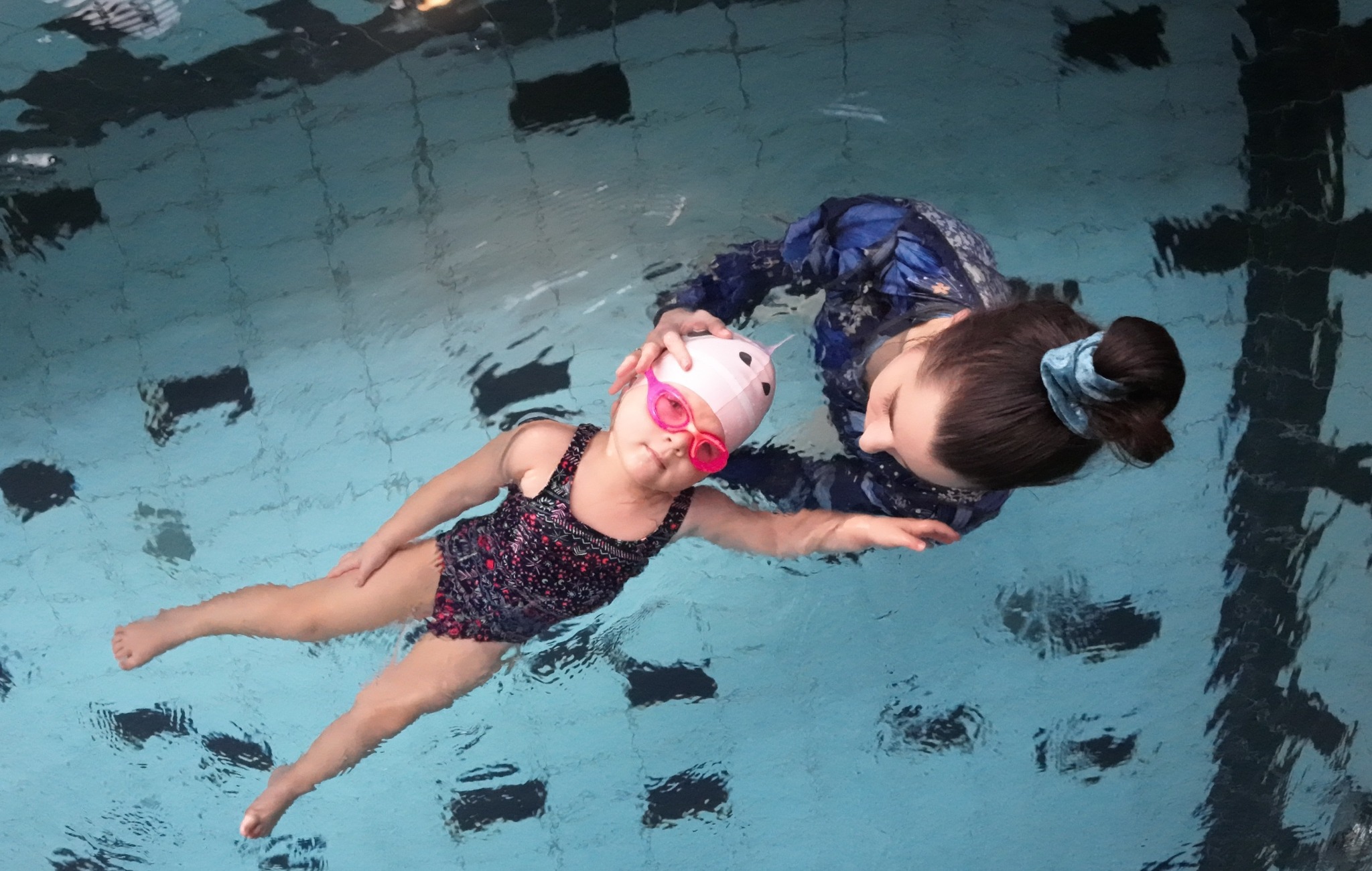 Dziewczynka w różowym czepku i okularach pływa w basenie z instruktorką, widok z góry, woda lekko wzburzona.