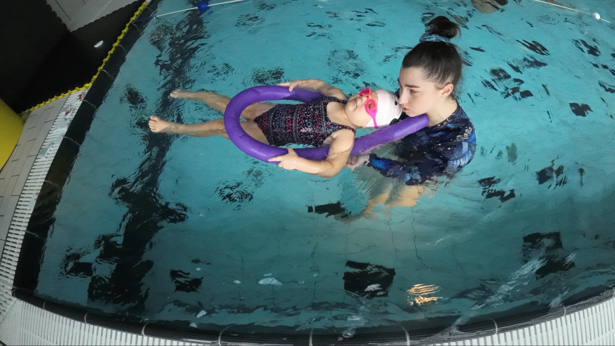 Instruktorka podtrzymuje małą dziewczynkę w różowych okularkach, unoszącą się na wodzie z fioletowym makaronem do pływania, widok z góry na basen.