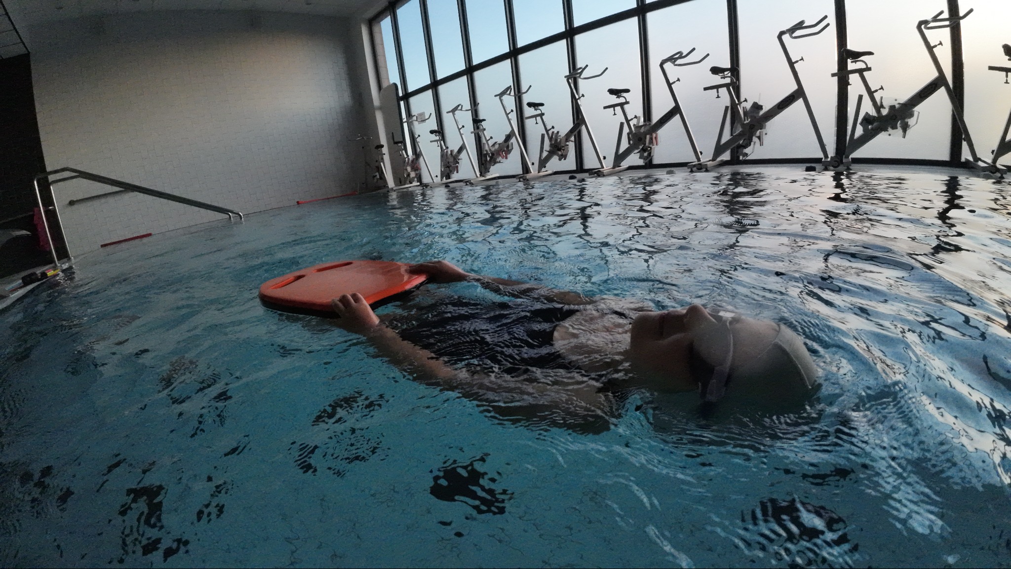 Osoba w czepku i okularach pływa na plecach w basenie, trzymając pomarańczową deskę; w tle okna i rowery wodne.