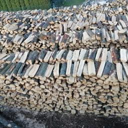 Stos pociętych i połupanych kawałków drewna ułożonych warstwami, widoczne sęki i kora, tło z zielenią drzew.