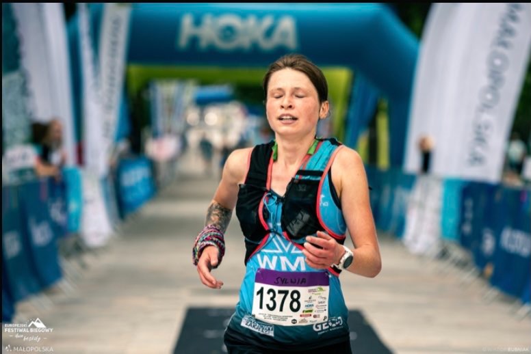 Kobieta biegnąca w zawodach, z numerem startowym 1378, zbliża się do mety, widoczne logo Hoka i banery reklamowe wydarzenia.