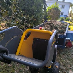 KNT Usługi leśno ogrodnicze Nikola Balcyrzak - Żółto-szara rębarka do gałęzi z przyczepą wypełnioną rozdrobnionym materiałem roślinnym na tle zieleni i białego budynku.