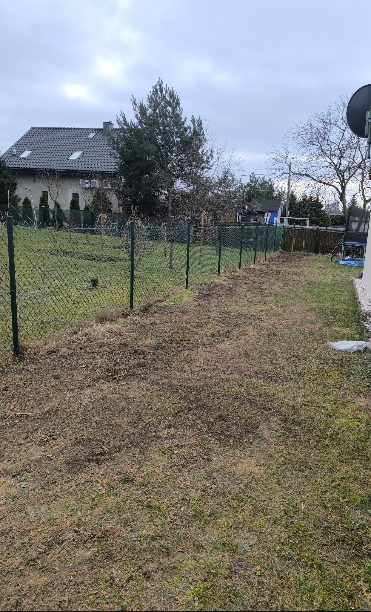 Długi, nowy, zielony płot z siatki oddzielający trawnik od ogrodu, z domem w tle