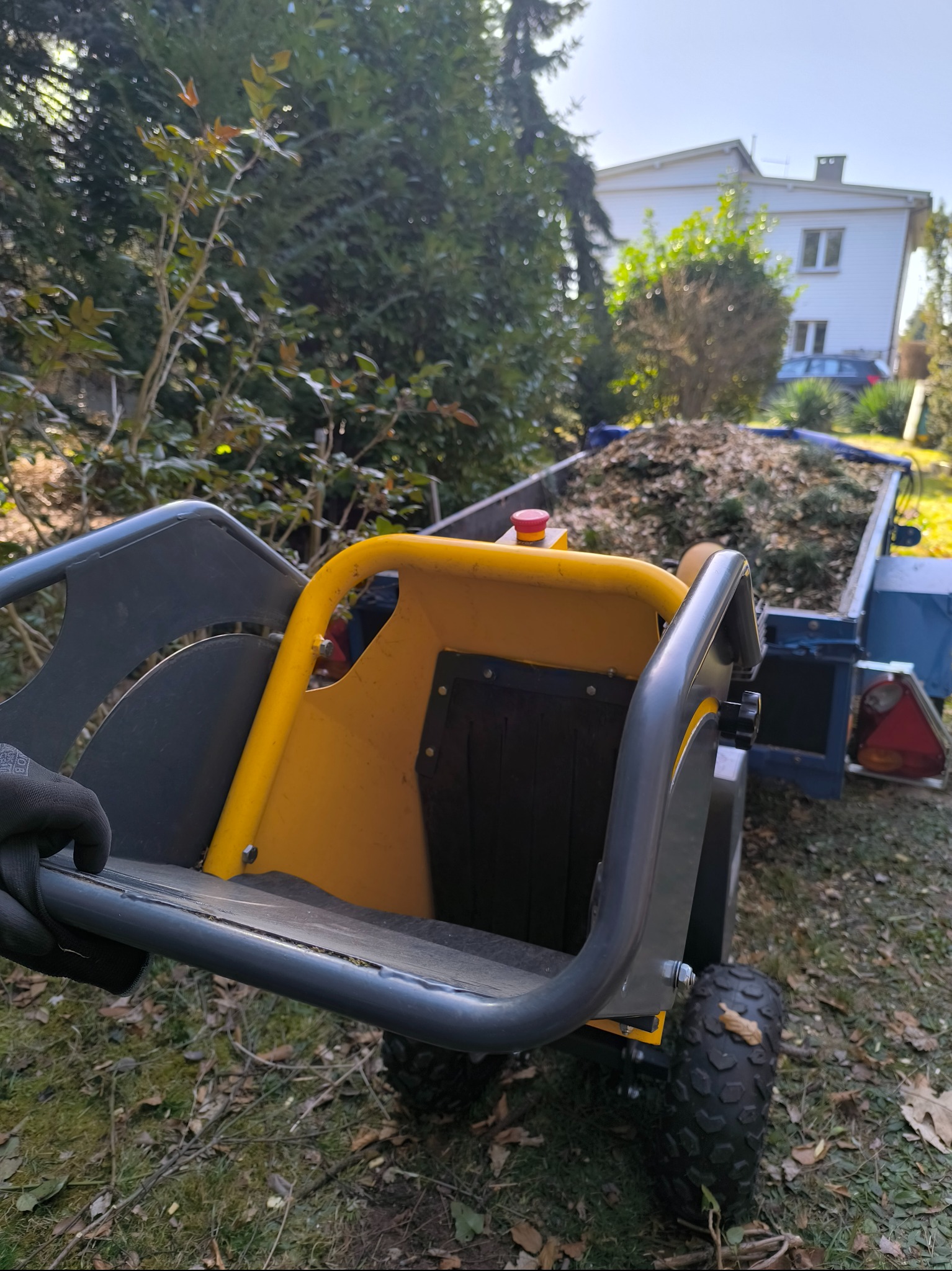 Żółto-szara rębarka do gałęzi z przyczepą wypełnioną rozdrobnionym materiałem roślinnym na tle zieleni i białego budynku.