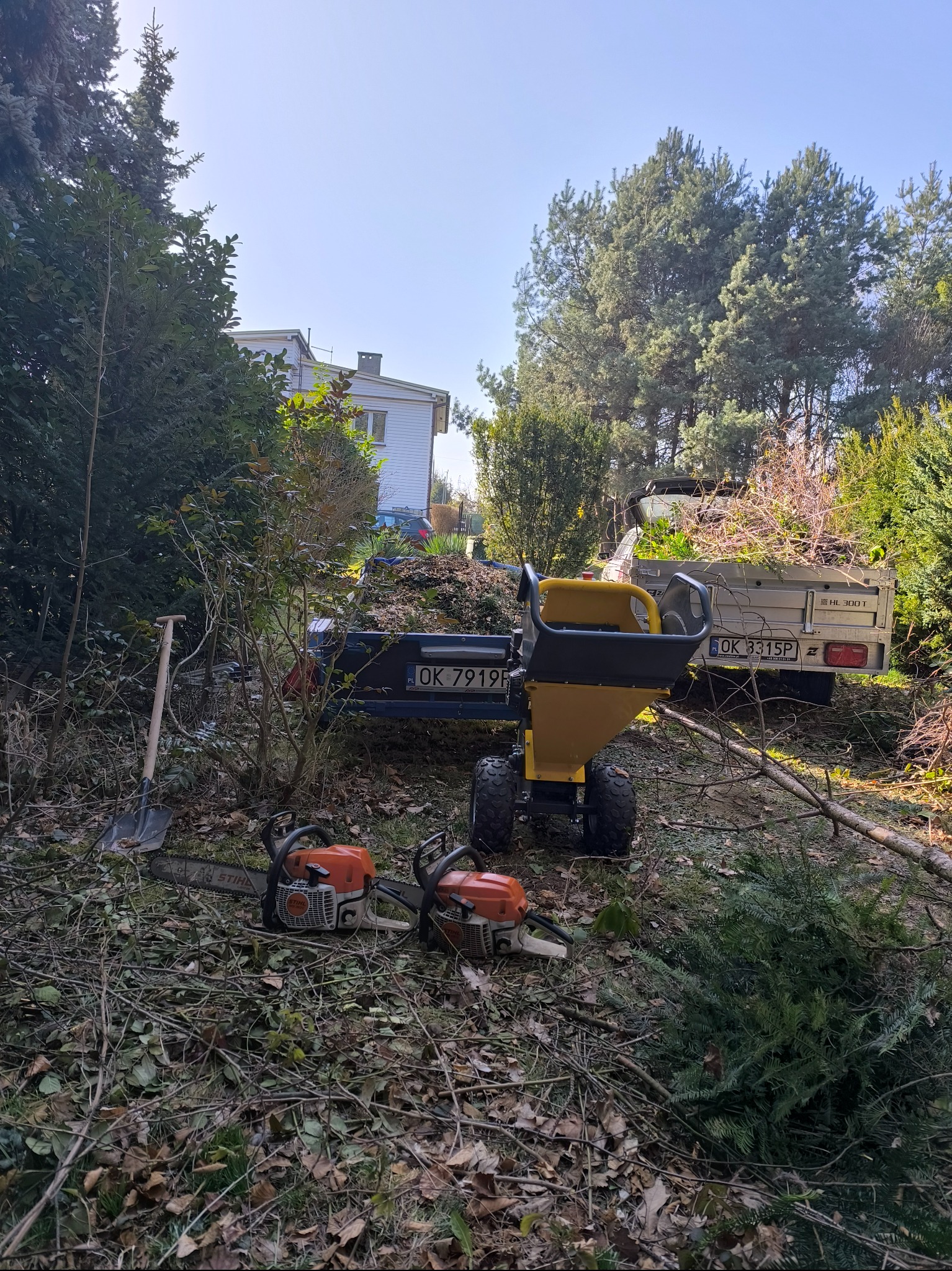 Dwie piły spalinowe Stihl leżące na ziemi obok stosu gałęzi i żółto-czarnego rębaka do gałęzi, z przyczepą wypełnioną rozdrobnionymi gałęziami w tle, widok z poziomu gruntu na tle drzew i domu.