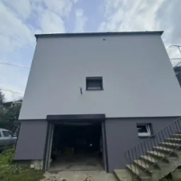 Dwukondygnacyjny dom z garażem i schodami zewnętrznymi, świeżo po remoncie elewacji w odcieniach szarości, z widocznym samochodem osobowym i fragmentem zieleni.