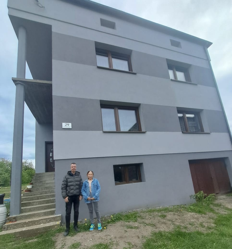 Trzy kondygnacyjny dom w odcieniach szarości z brązowymi oknami. Przed budynkiem stoją dwie osoby. Widoczny numer domu 29 i garaż z brązową bramą.