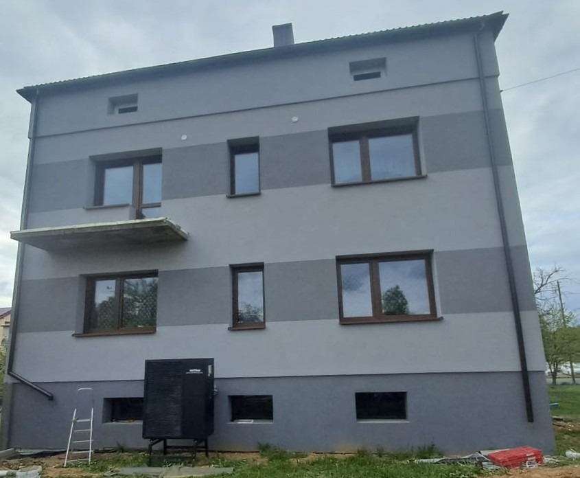 Odnowiona elewacja dwukondygnacyjnego budynku z szarymi pasami, nowymi oknami i zamontowaną pompą ciepła przy ścianie.  Drabina oparta o ścianę.