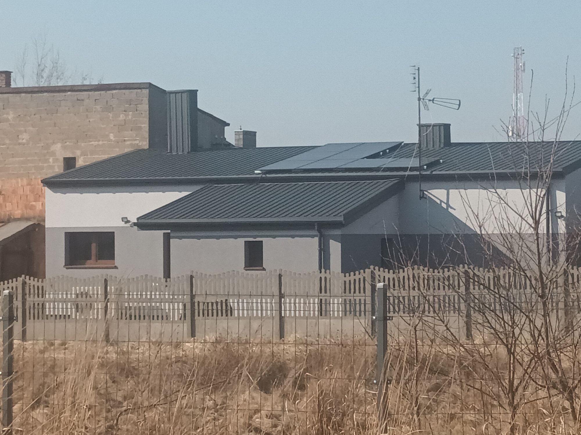 Dom jednorodzinny z panelami słonecznymi na dachu, widziany zza ogrodzenia. Szara elewacja i grafitowa blacha dachowa. Antena telewizyjna na kominie.