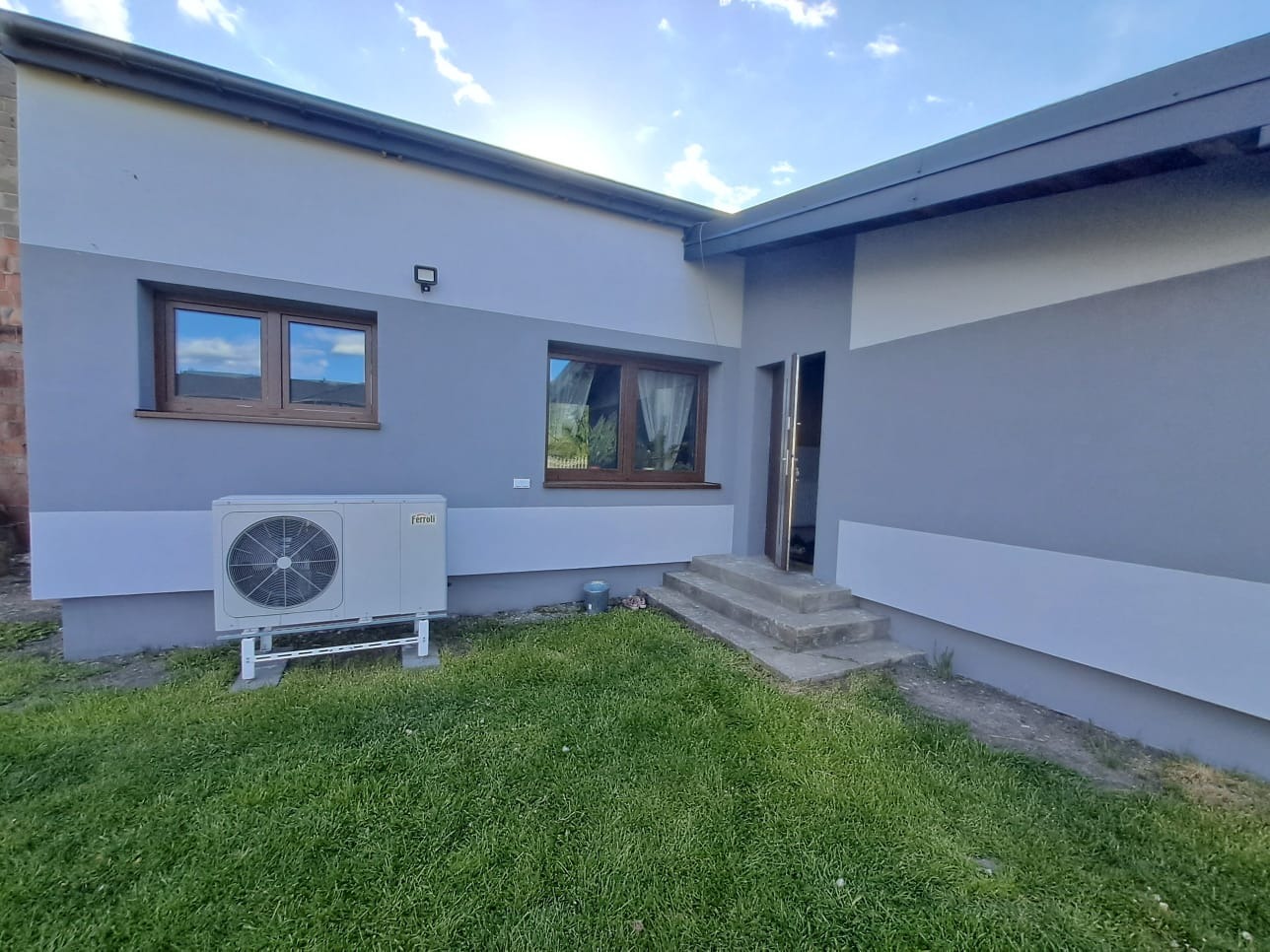 Zamontowana jednostka zewnętrzna pompy ciepła Ferroli przy szarym budynku z brązowymi oknami na tle zielonego trawnika w słoneczny dzień.
