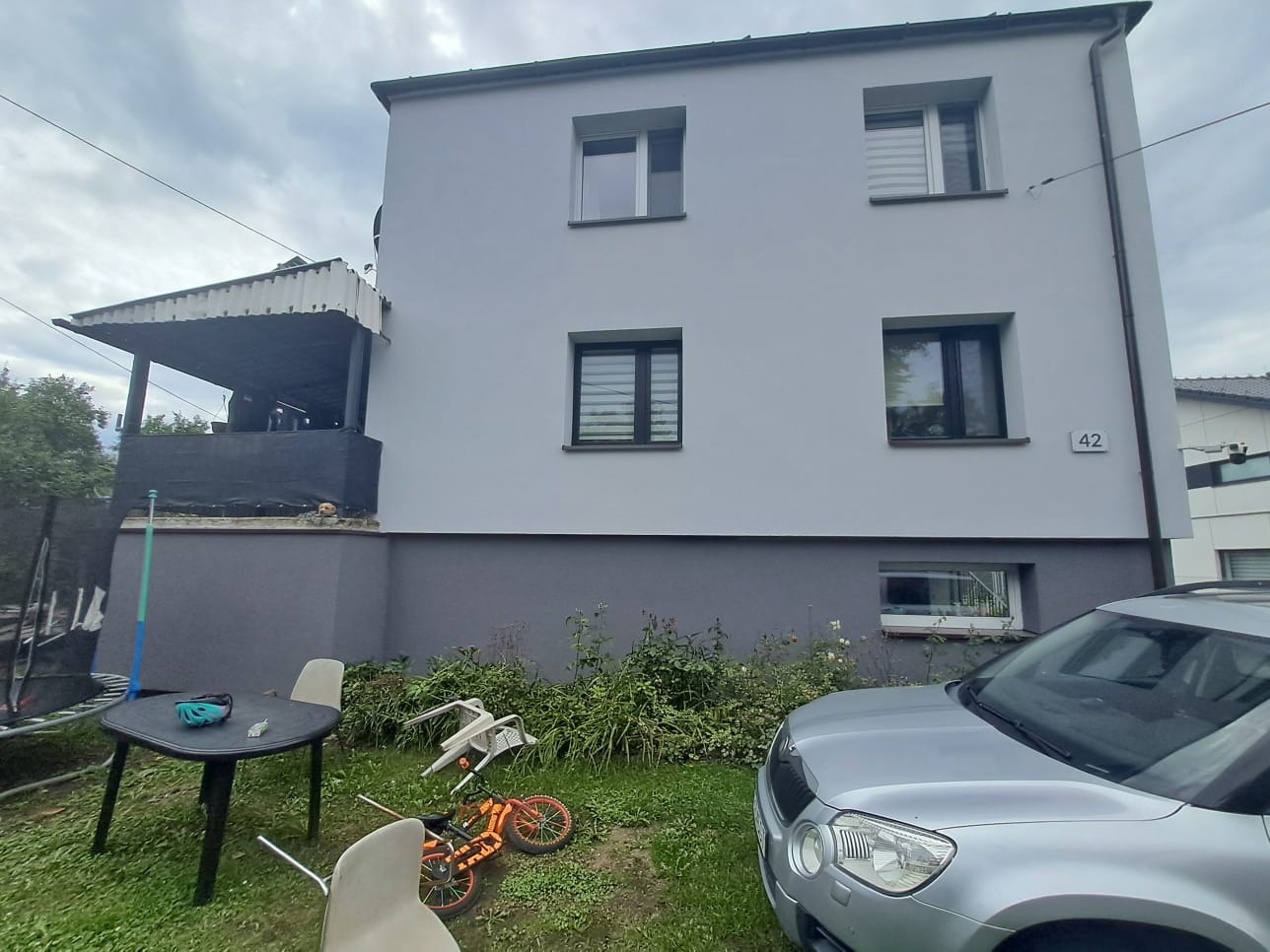 Dwu kondygnacyjny dom z szarą elewacją, widoczny samochód, ogród z meblami i zabawkami. Dom po remoncie elewacji.