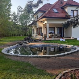 ARTEX Artur Rączka - Oczko wodne z kamiennym obrzeżem w trakcie budowy, widoczna folia zabezpieczająca, w tle dom jednorodzinny z tarasem i zadbany trawnik.