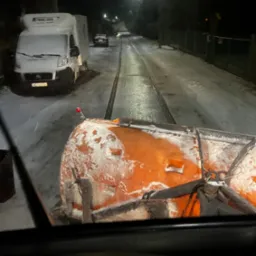 Widok z kabiny na odśnieżarkę w trakcie pracy na ośnieżonej ulicy nocą, z widocznym białym vanem i innymi pojazdami w oddali.