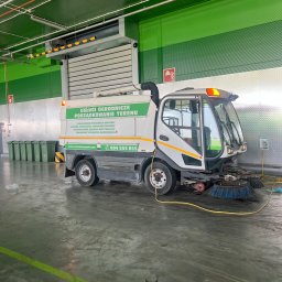 ARTEX Artur Rączka - Maszyna do zamiatania z napisem 'Usługi ogrodnicze porządkowanie terenu' stoi w hali, widoczne szczotki zamiatające i pojemniki na śmieci w tle.