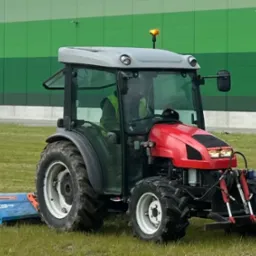 Czerwony traktor z kabiną i zamontowaną kosiarką rotacyjną na trawniku przed nowoczesnym budynkiem w odcieniach zieleni.