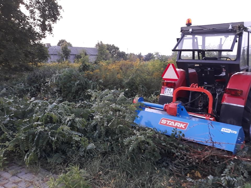 Czerwony traktor z niebieskim mulczerem Stark KHM 175 tnący zarośla na zaniedbanym terenie, w tle widoczny budynek gospodarczy.