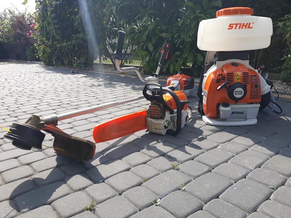 Zestaw sprzętu ogrodniczego marki Stihl: kosa spalinowa z osłoną, podkaszarka i opryskiwacz plecakowy, ułożone na szarej kostce brukowej, w tle zielone krzewy.