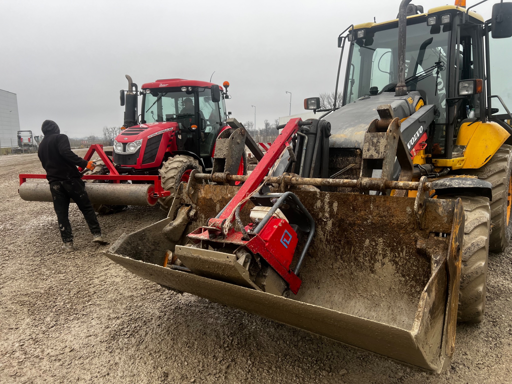 Czerwony traktor z wałem ugniatającym i żółta ładowarka Volvo z ubijarką w łyżce, na żwirowym placu, operator w czarnej kurtce przy traktorze.