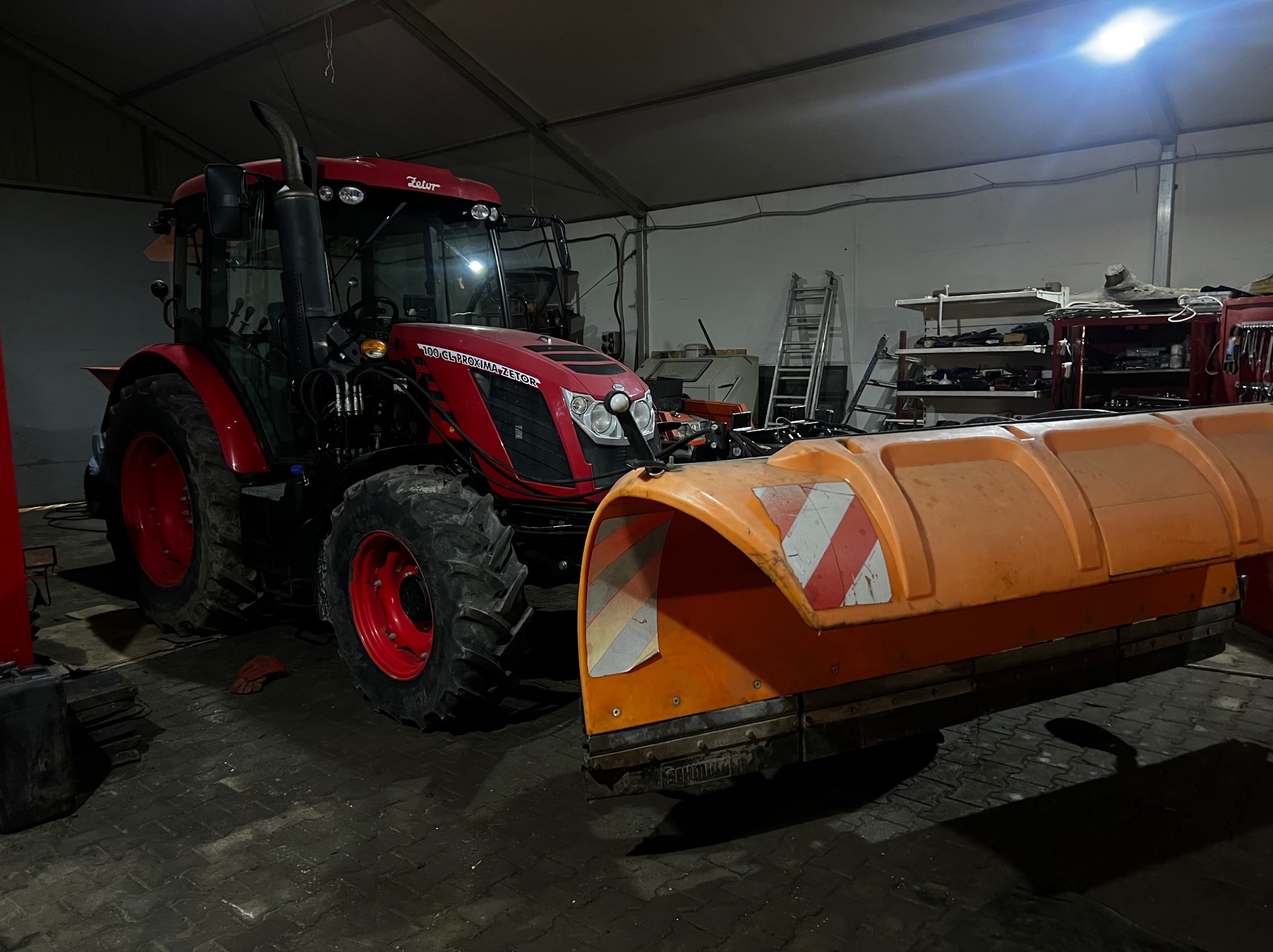 Czerwony traktor Zetor z zamontowanym pomarańczowym pługiem śnieżnym w garażu, widoczne półki z narzędziami i drabina.
