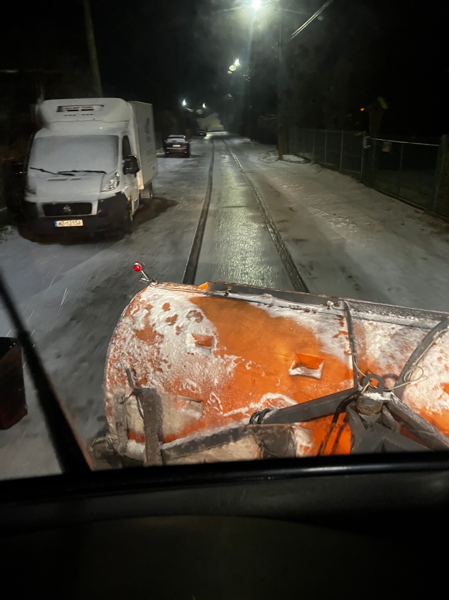 Widok z kabiny na odśnieżarkę w trakcie pracy na ośnieżonej ulicy nocą, z widocznym białym vanem i innymi pojazdami w oddali.